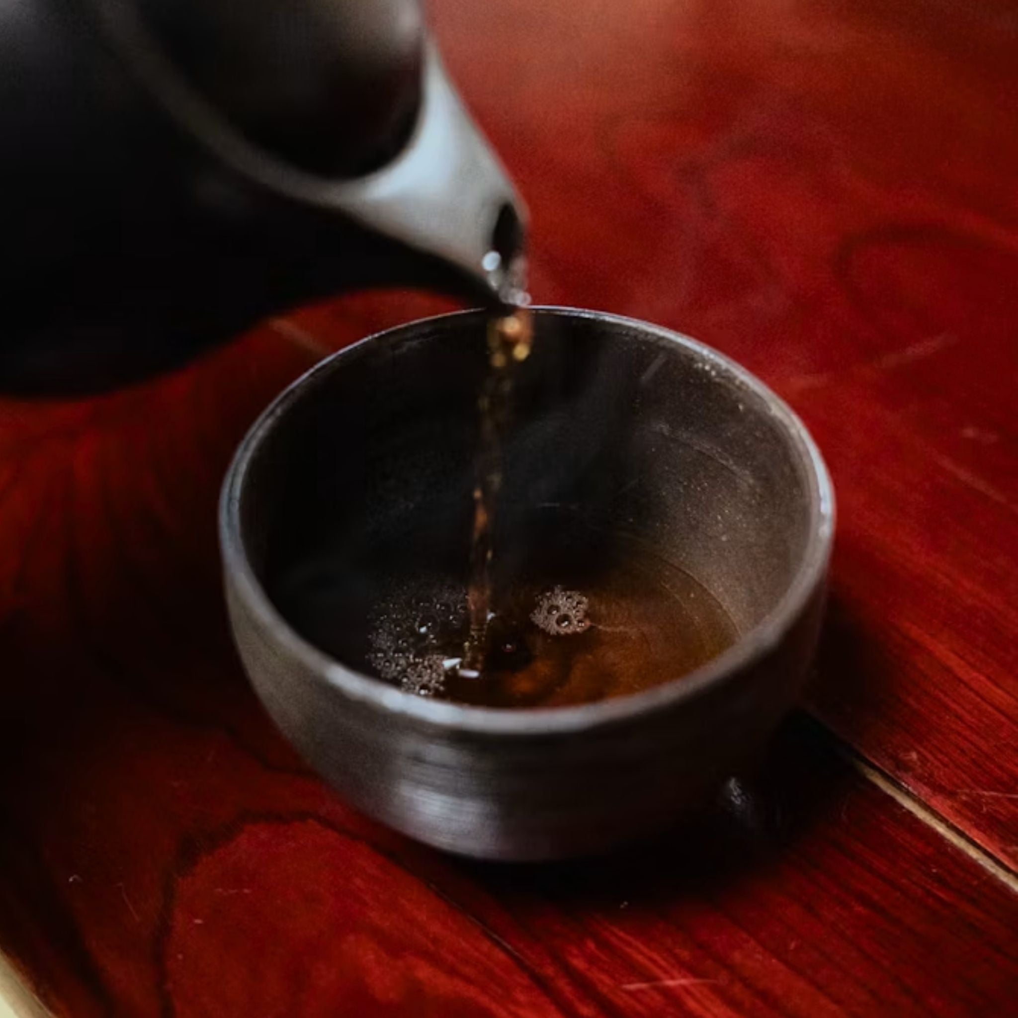 Dark ceramic tea cup on a wooden table with tea being poured from a black teapot.