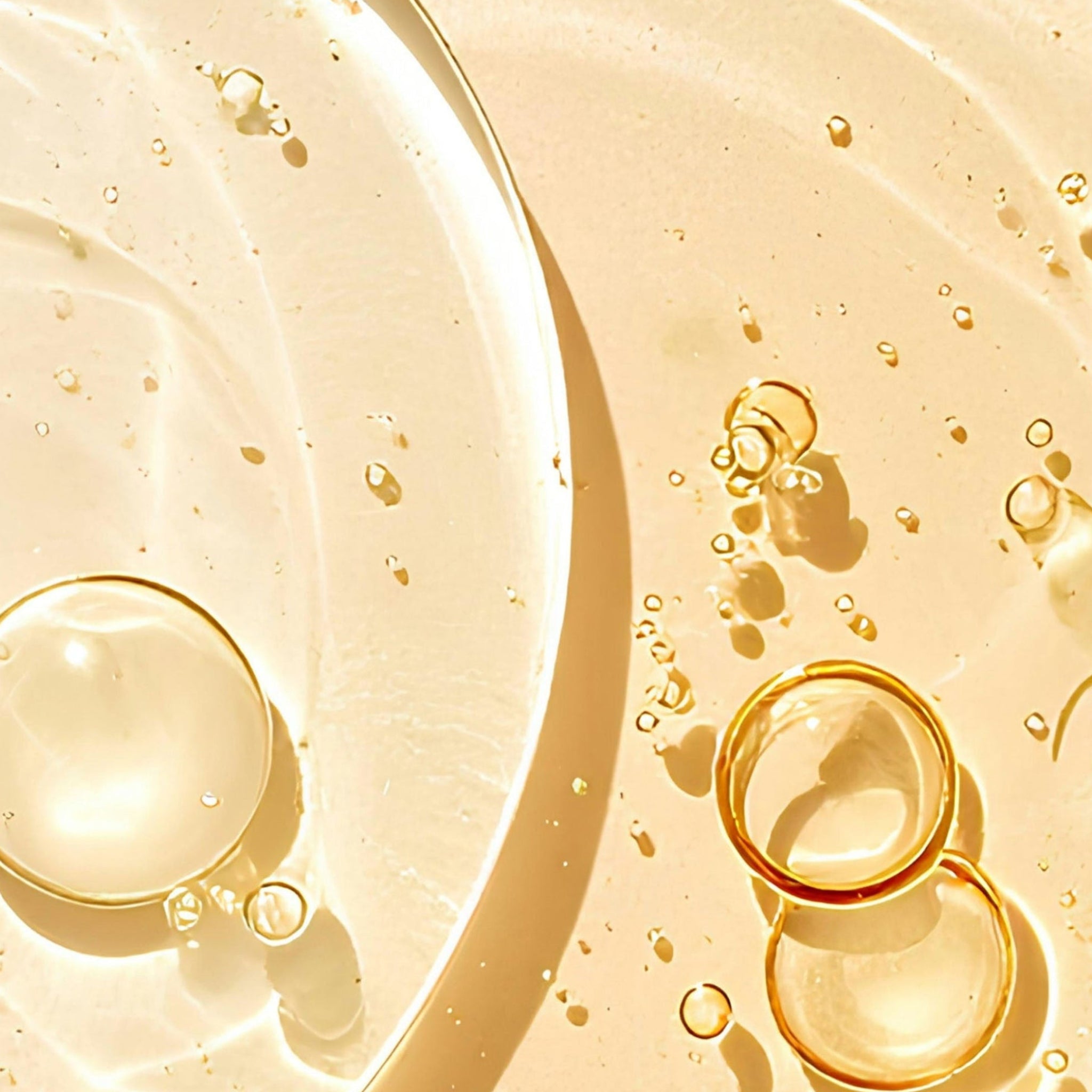 Close-up of a glossy, creamy golden-beige cosmetic cream with translucent oil droplets and bubbles on a warm beige surface.