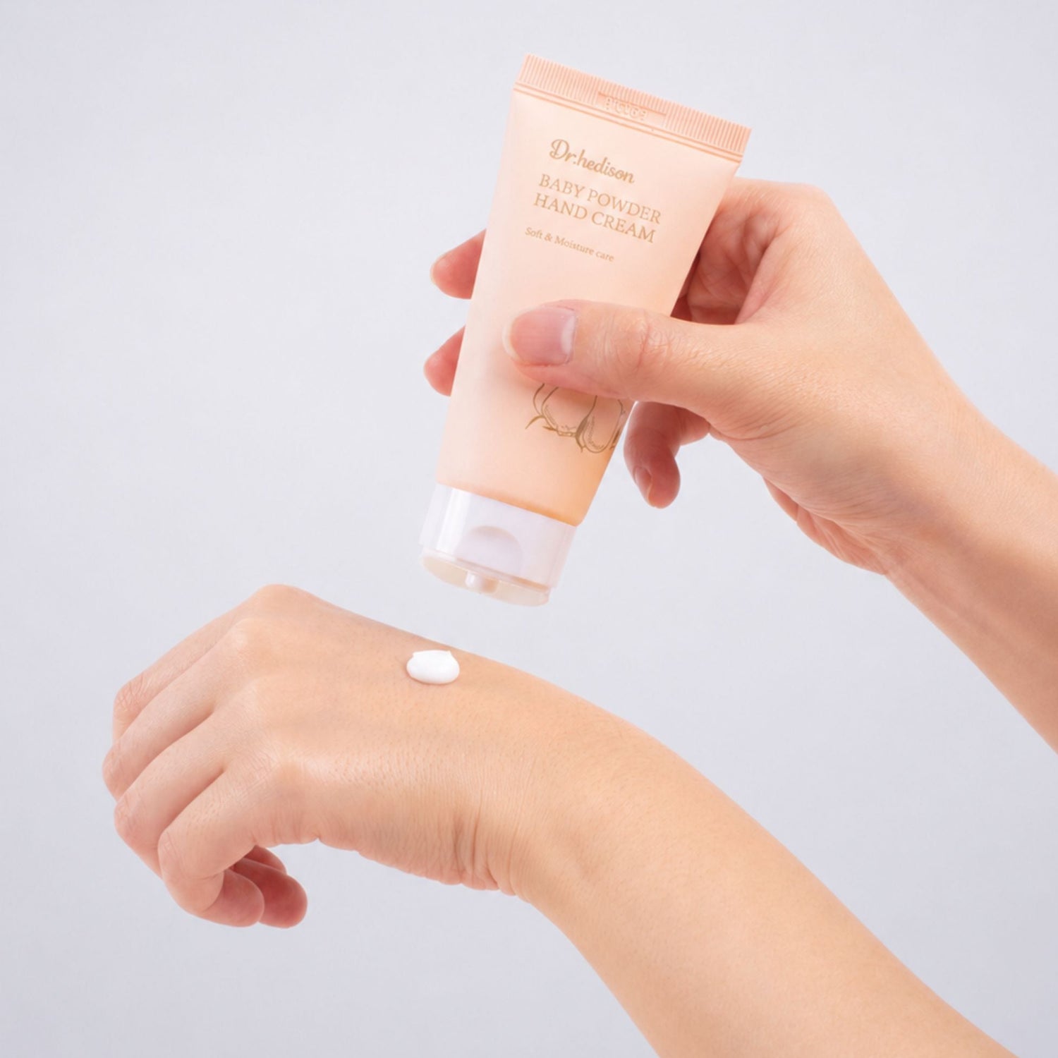 A hand holds a squeeze tube of Hand Cream above another hand showing a small dollop of the white cream, set against a clean light gray background.